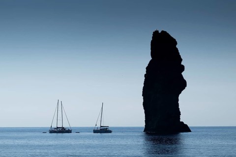 Silhouette of a towering monumental rock pin, sailboats in the calm sea, near Filicudi, Aeolian or