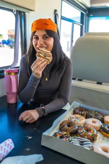 Young woman wearing a bandana and long sleeve shirt biting into a glazed donut from a large box of