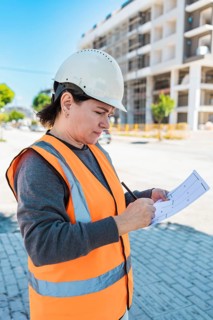 Woman wearing a hard hat and safety vest inspects a construction site. She holds a clipboard and