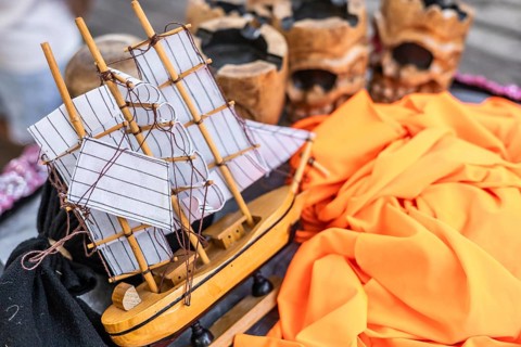 A detailed model ship with white sails rests on a colorful fabric background. Decorative skulls are
