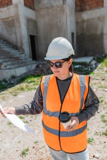 Woman in a hard hat and safety vest holds a coffee cup and construction plans at a building site.