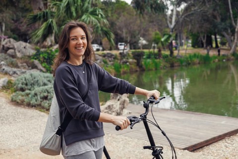 Smiling young Caucasian woman with brown hair stands beside a bicycle near a serene lake in a park.
