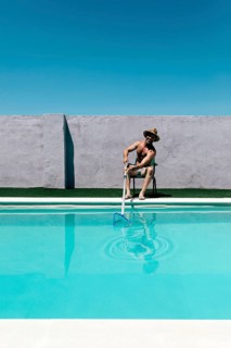 A shirtless man in shorts and a hat cleans a swimming pool with a long net skimmer under a bright