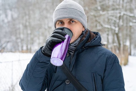 A man in winter attire drinking from a purple thermos on a snowy day. A forty-year-old man stands