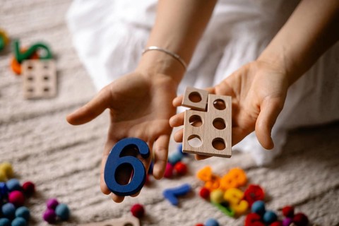 Hands holding a blue number six and wooden toy with colorful scattered toys
