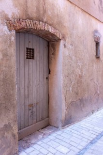 A weathered wooden door sits beneath a brick arch in a textured pink wall beside stone paving,