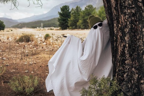 A playful ghost figure draped in a white sheet with sunglasses, leaning against a tree in a rural