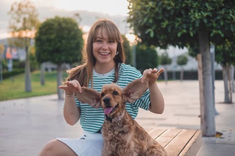A woman playfully holds a dog's ears while sitting on a bench in a sunlit park