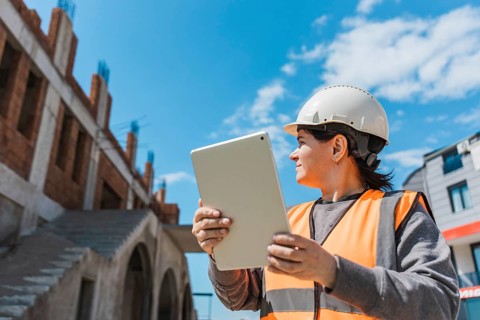Woman in a hard hat and safety vest holds a tablet at a construction site. The building is in