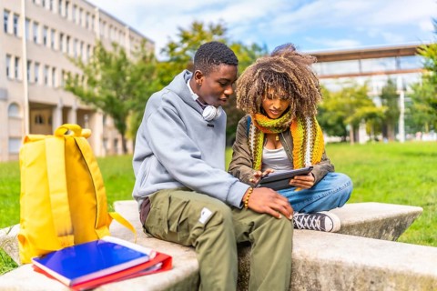 Friends using tablet to study together sitting outside the university campus in a sunny day of