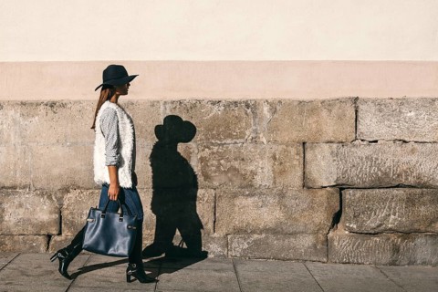 A fashionable woman wearing a hat, sweater, and boots walks by a stone wall. Her stylish ensemble