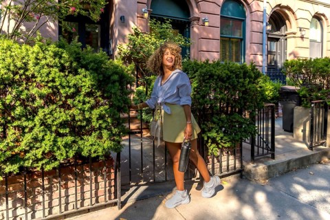 Woman with curly hair smiling and carrying a reusable bottle, walking out of a brownstone's gated