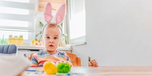 Banner with child wearing bunny ears sits at a table decorated for Easter. Colorful eggs are placed