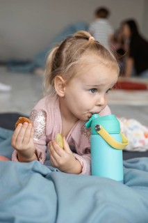 A young Caucasian girl with blonde hair enjoys snacks while holding a blue water bottle. The scene