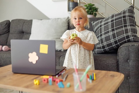 Young blond European girl standing near a laptop on a wooden table with colorful toys in a living