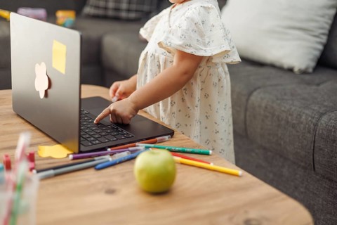 Young toddler girl with light brown hair in white dress using laptop on wooden table with colored