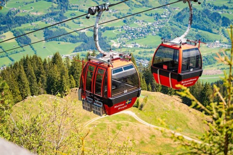 Two red cable car cabins float above a wooded hilly landscape with a wide view, Hahnenkamm,