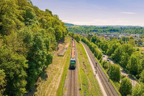 Green, hilly landscape with rails and a train, lined with trees and vegetation, electrification of