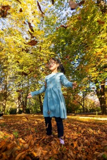 A joyful Asian child in a blue dress happily plays amid falling autumn leaves in a vibrant park