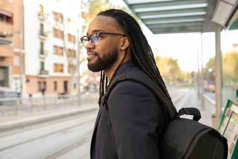 A cuban business man with dreadlocks and glasses waits patiently at a tram stop. He carries a