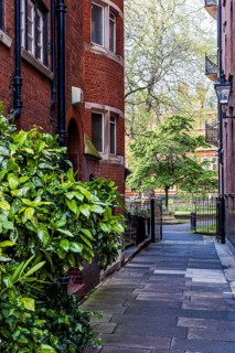 A paved lane flanked by red brick buildings leads to the entrance of Mount Street Gardens in
