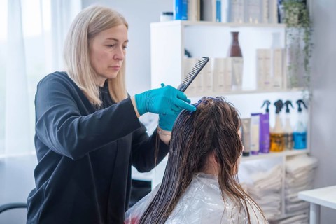 A hair stylist performs a keratin hair straightening treatment. The client sits in a salon chair