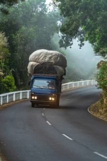 Blue truck carrying a large, canvas covered load of cargo along a winding road through a misty,