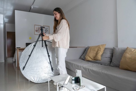 Side view of woman adjusting camera tripod legs in living room, with silver reflector and phone