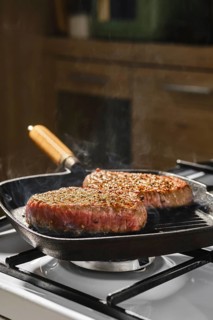 Two beef steaks are cooking on cast iron grill pan on gas stove in kitchen. Steam rises from meat