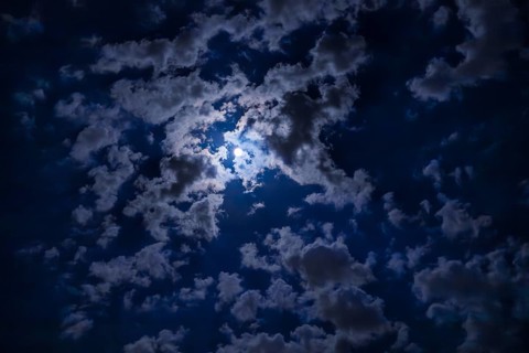 A night sky filled with dark clouds partially illuminated by a bright moon. The scene captures the