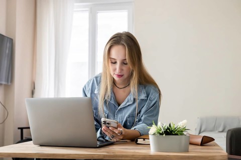 Young Caucasian woman with long blonde hair using a smartphone while working on a laptop at a