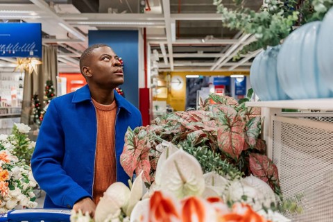A young Black man with short hair wearing a blue coat and orange sweater looks up while shopping in