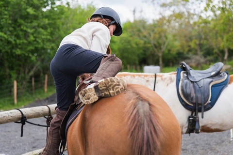 A young girl in a helmet and riding boots mounts a horse on a farm. The scene captures a fun and
