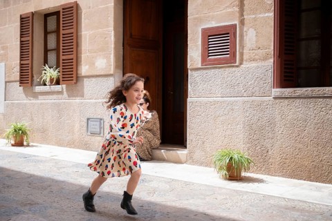 A young girl joyfully runs through a sunlit village street, her patterned dress fluttering as she