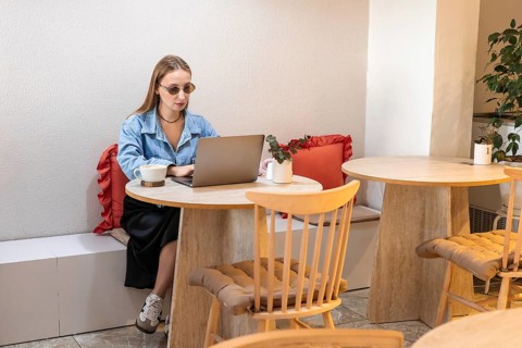 A young Caucasian woman with long blonde hair works on a laptop at a cafe table. She wears