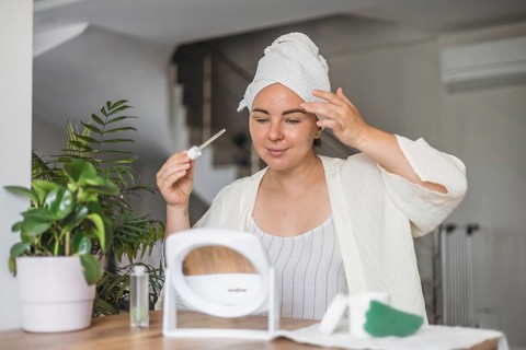 A young Caucasian woman with brown hair in a towel applies serum to her face while preparing for a