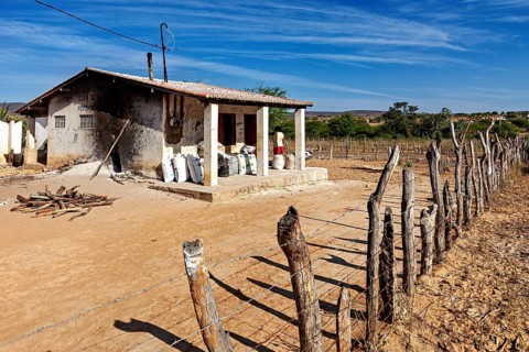 A small house in a dry landscape with a simple wooden fence in the foreground, a cassava mill in