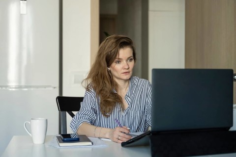 Woman in striped shirt works at home, focused on laptop. She writes in a notebook with a cup on the