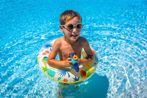 Boy aims with a water gun while playing with a float inside a blue swimming pool in summer