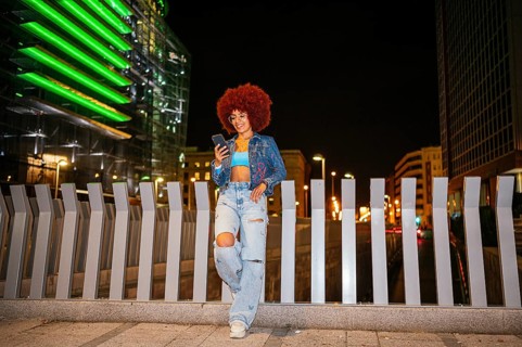 An African descendent woman with vibrant afro hair stands by a city barricade while using her