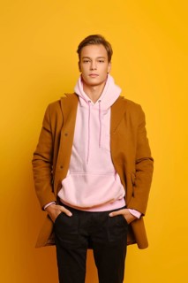 Handsome stylish man in brown coat and pink hoodie sweater posing with hands in pockets over yellow