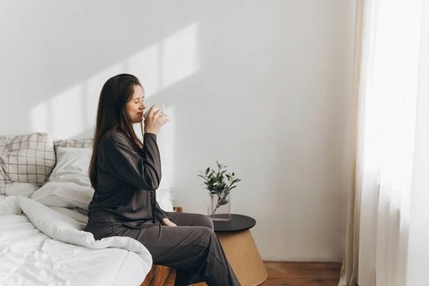 A woman in comfortable loungewear enjoys a glass of water by her bed in a sunlit room, promoting