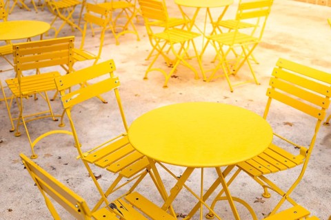 Rows of bright yellow metal tables and chairs are arranged in an outdoor area. The sunny