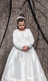 A young girl stands against a tree wearing a beautiful white First Communion dress, complete with a