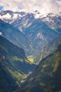 A wide valley stretches between high, partly snow-covered mountains under a blue sky, Stilluptal,