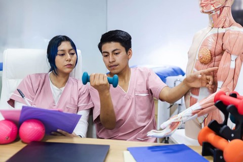 Two physiotherapy students in an Ecuador clinic study anatomy with a muscular model and weights,
