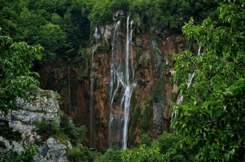 A mighty, narrow waterfall falls along a rock wall surrounded by green forest, Plitvice Lakes