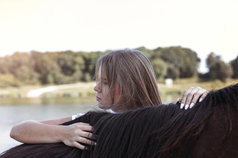 A young girl enjoys a peaceful moment with a horse by the lakeside. The serene setting features a