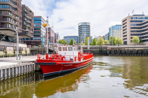 A red boat is lying in the city harbor surrounded by modern buildings and mirrored water,