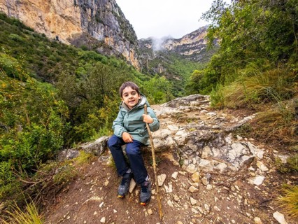 Young boy resting on a rocky mountain trail, holding a stick and smiling, surrounded by lush green